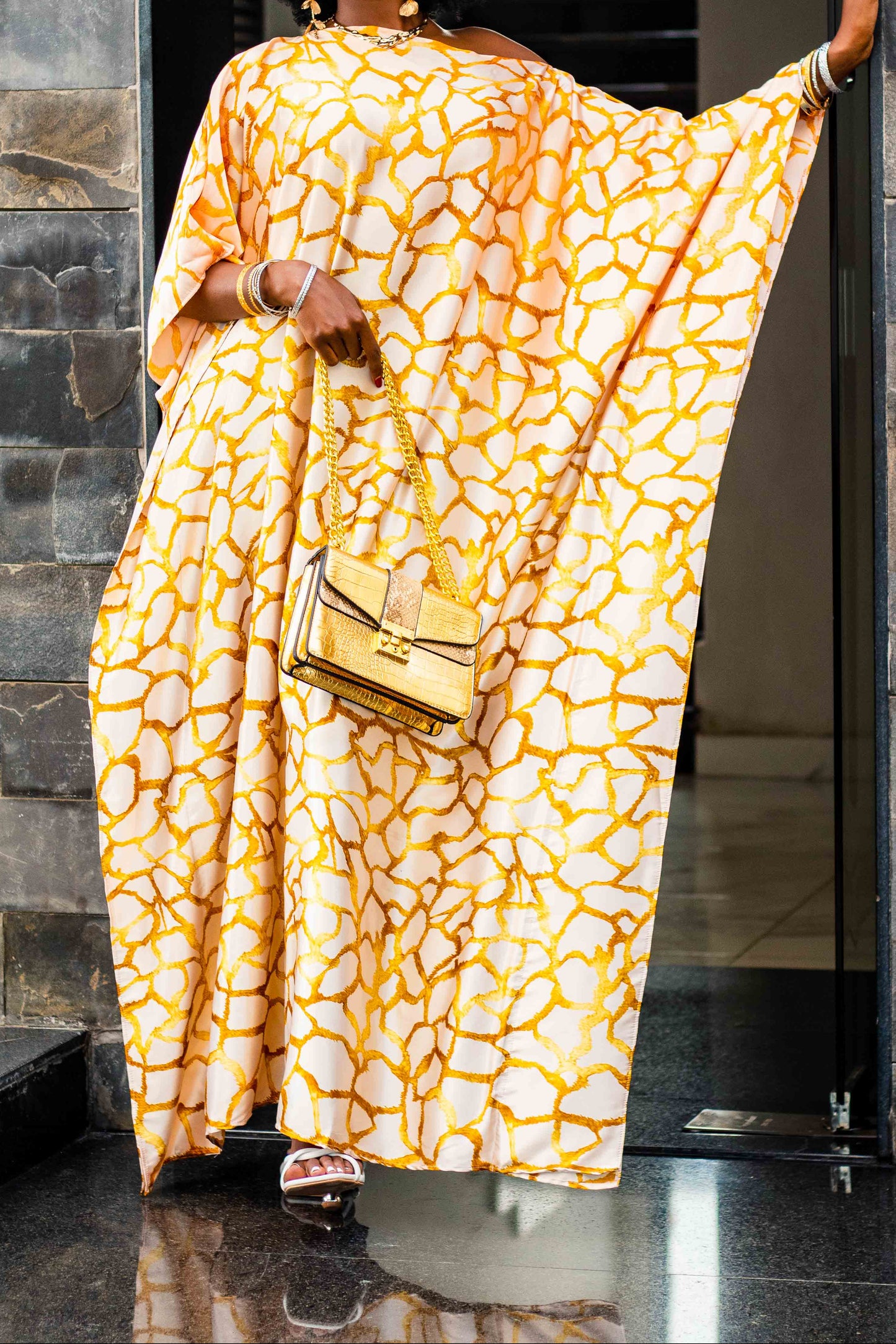 A woman with an afro hairstyle models a yellow and white abstract patterned kaftan dress, holding a matching yellow handbag, standing in a doorway with stone walls.