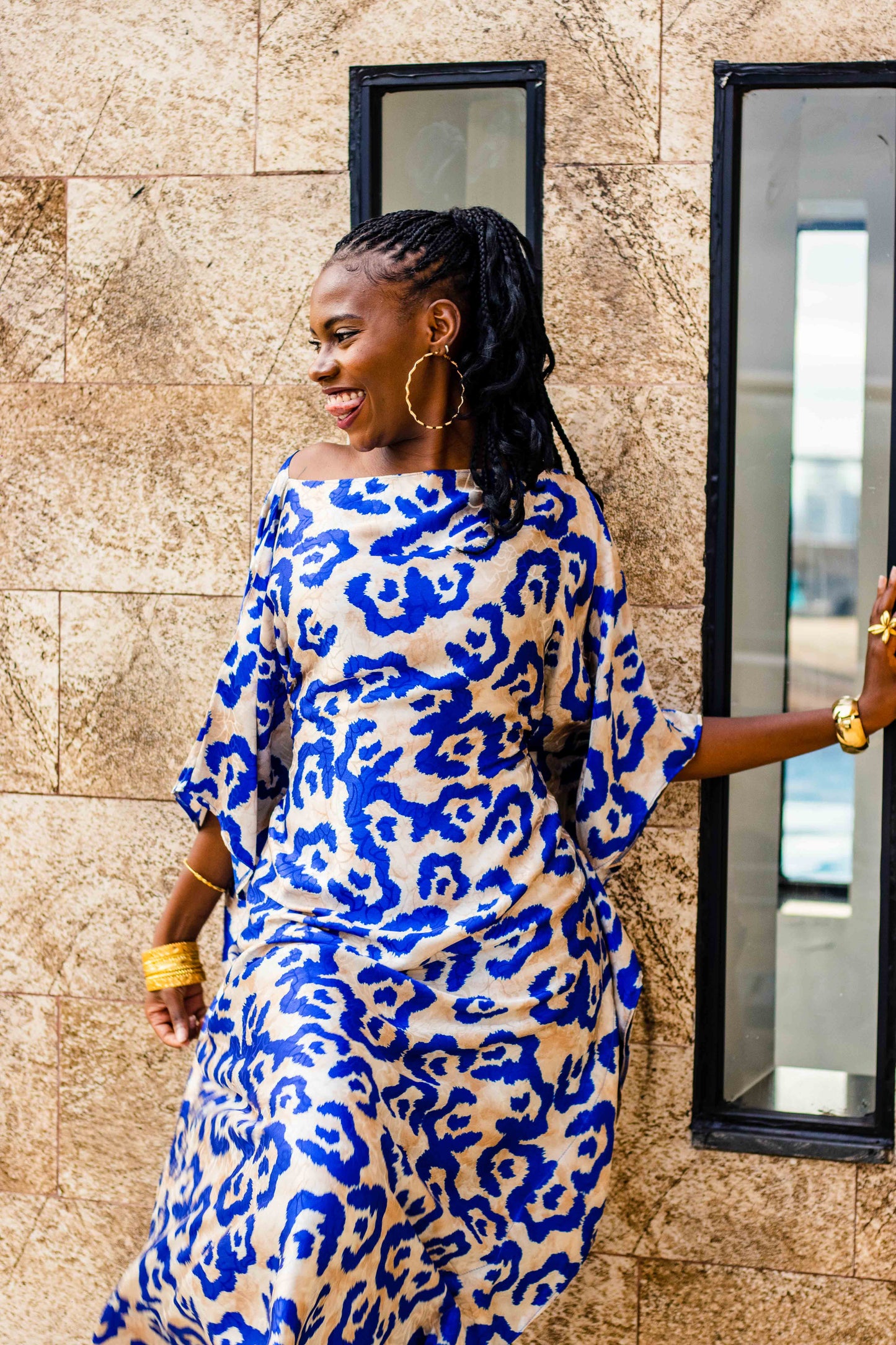 A woman wearing a blue and beige animal print dress leans against a stone wall next to a window.