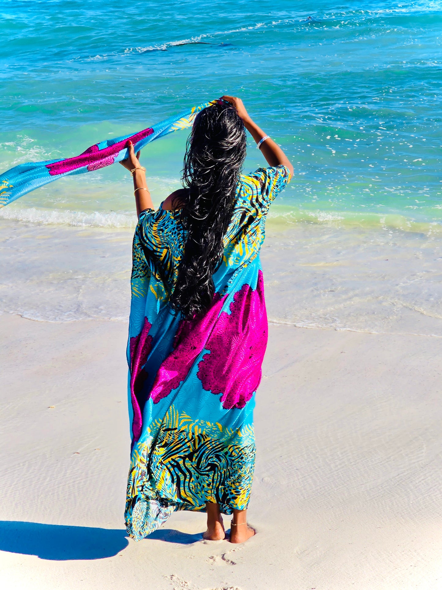 A woman stands on a sandy beach facing the ocean, wearing a flowing kaftan dress with a bold blue, pink, yellow, and black pattern. She holds a matching patterned scarf in the air with both hands.
