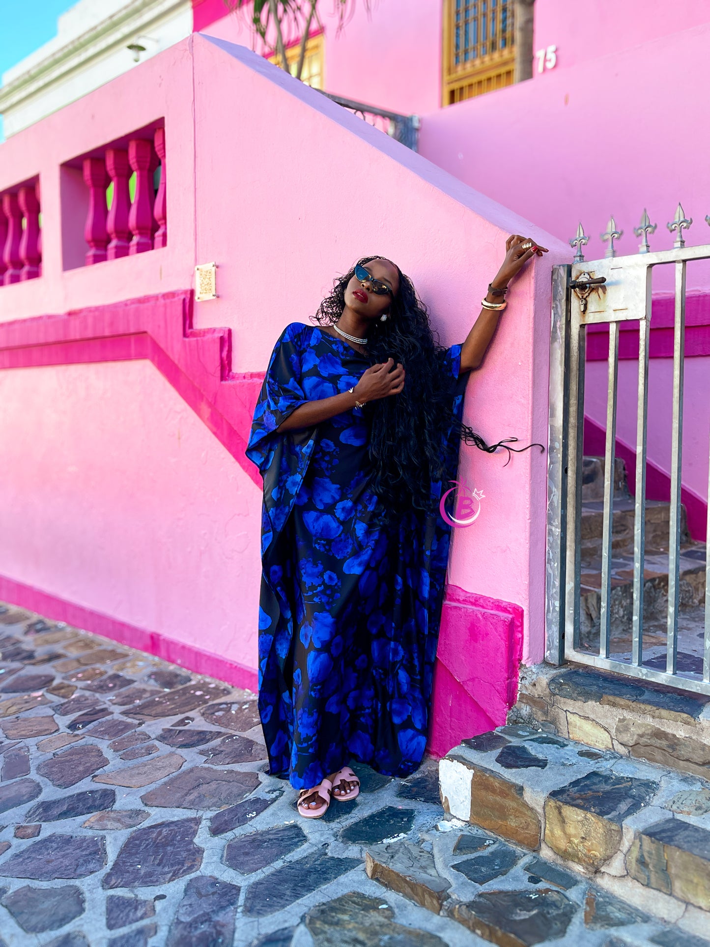 A woman wearing a blue and black patterned kaftan dress leans against a bright pink wall on a cobblestone street in Cape town.
