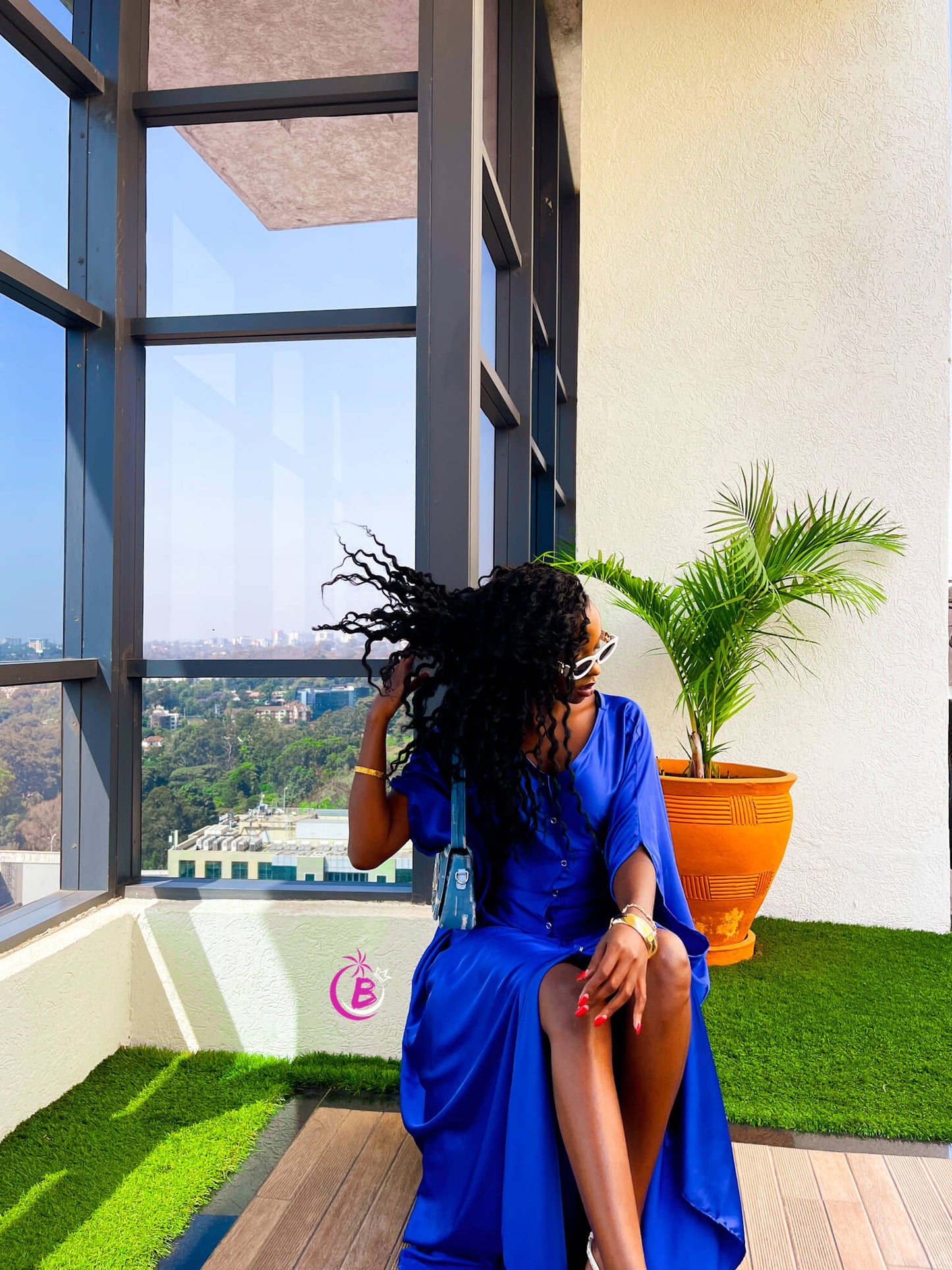 A woman wearing a vibrant royal blue dress and sunglasses sits on a wooden deck on a balcony with a view of a city skyline. She is next to a potted palm plant in an orange pot.