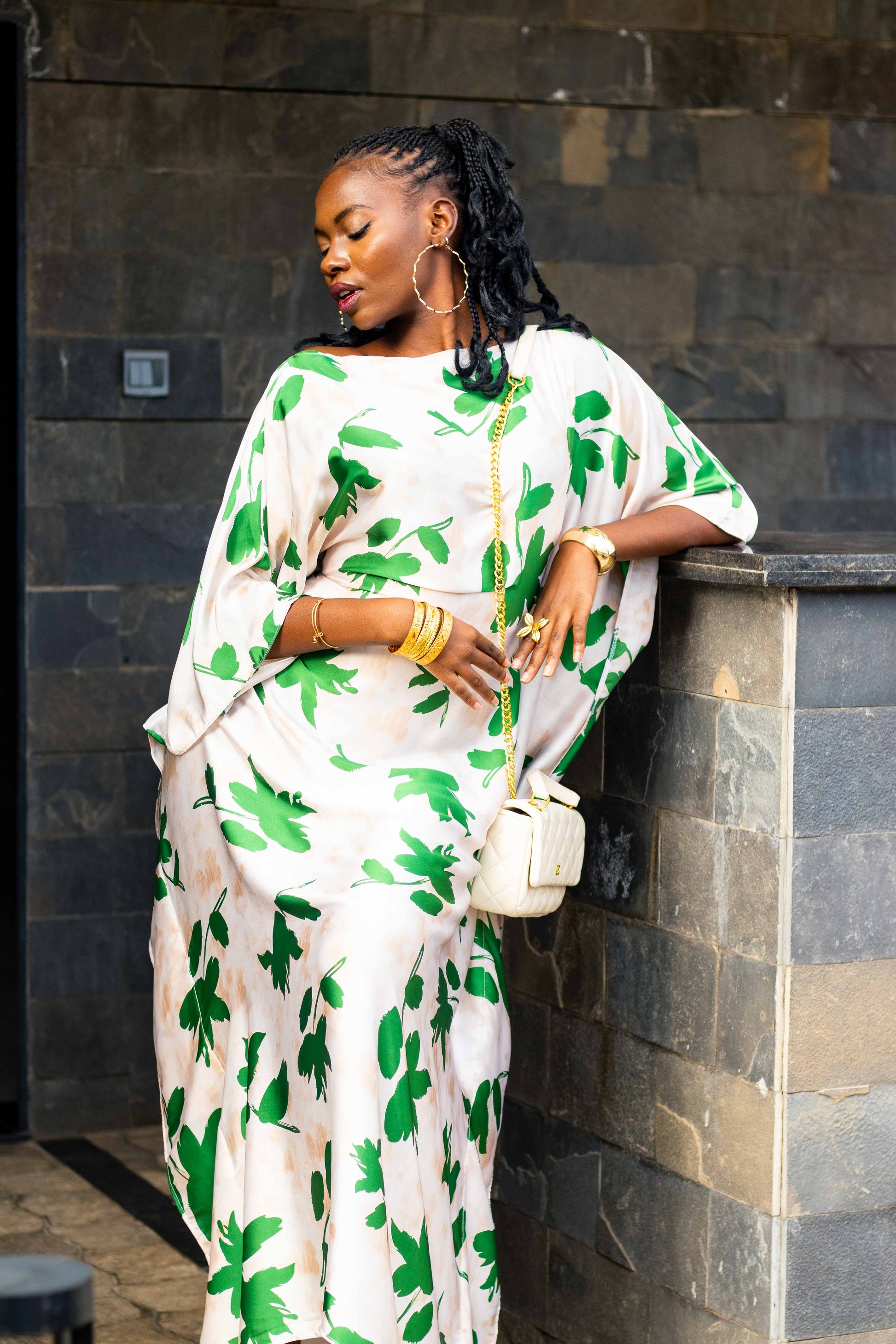 A woman wearing a white kaftan dress with a green leaf pattern, leaning against a dark stone wall. She is holding a small white handbag and wearing gold jewelry stacks.