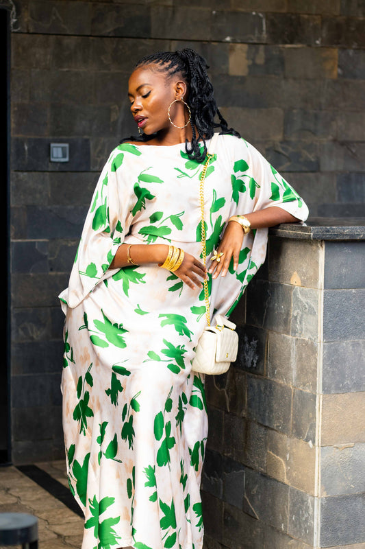 A woman wearing a white kaftan dress with a green leaf pattern, leaning against a dark stone wall. She is holding a small white handbag and wearing gold jewelry stacks.