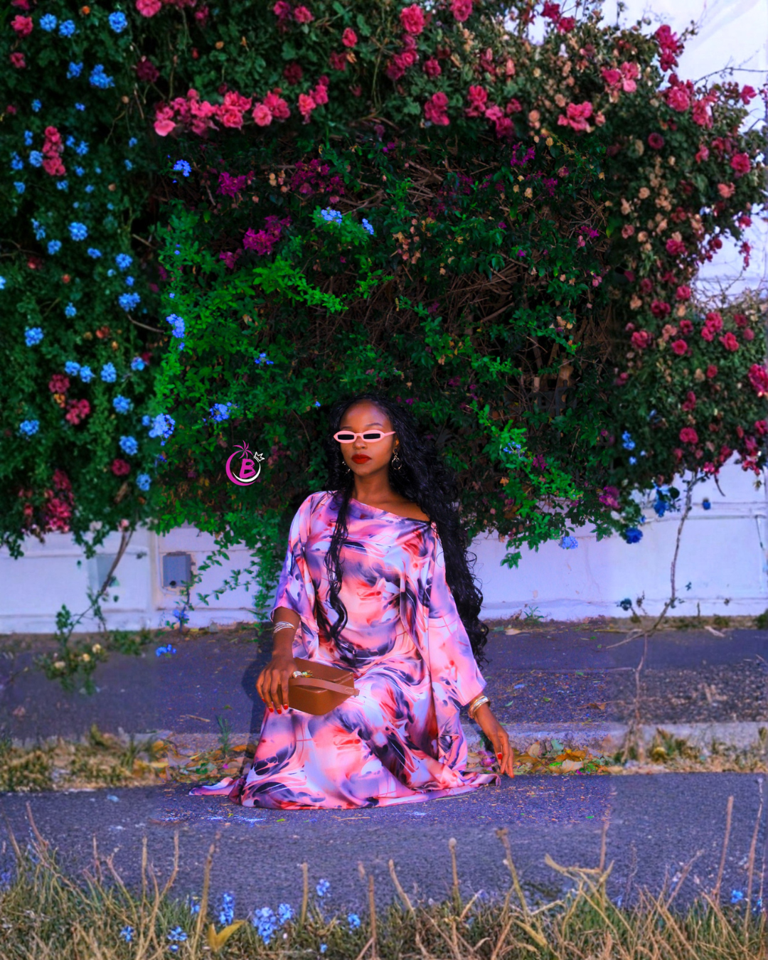 A woman wearing a pink and purple floral off-the-shoulder maxi dress sits on a paved path in front of a large flowering bush with pink and blue flowers.
