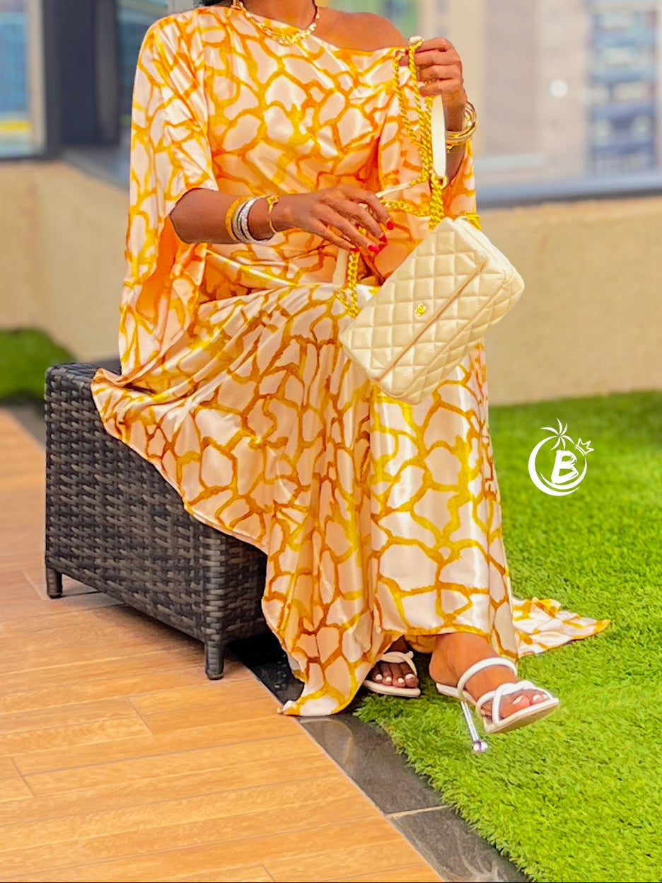 A woman with a large black afro sits on a dark wicker chair on a balcony, wearing a yellow and white patterned off-the-shoulder dress, holding a cream quilted handbag. A large potted palm is next to her, with a city view in the background.